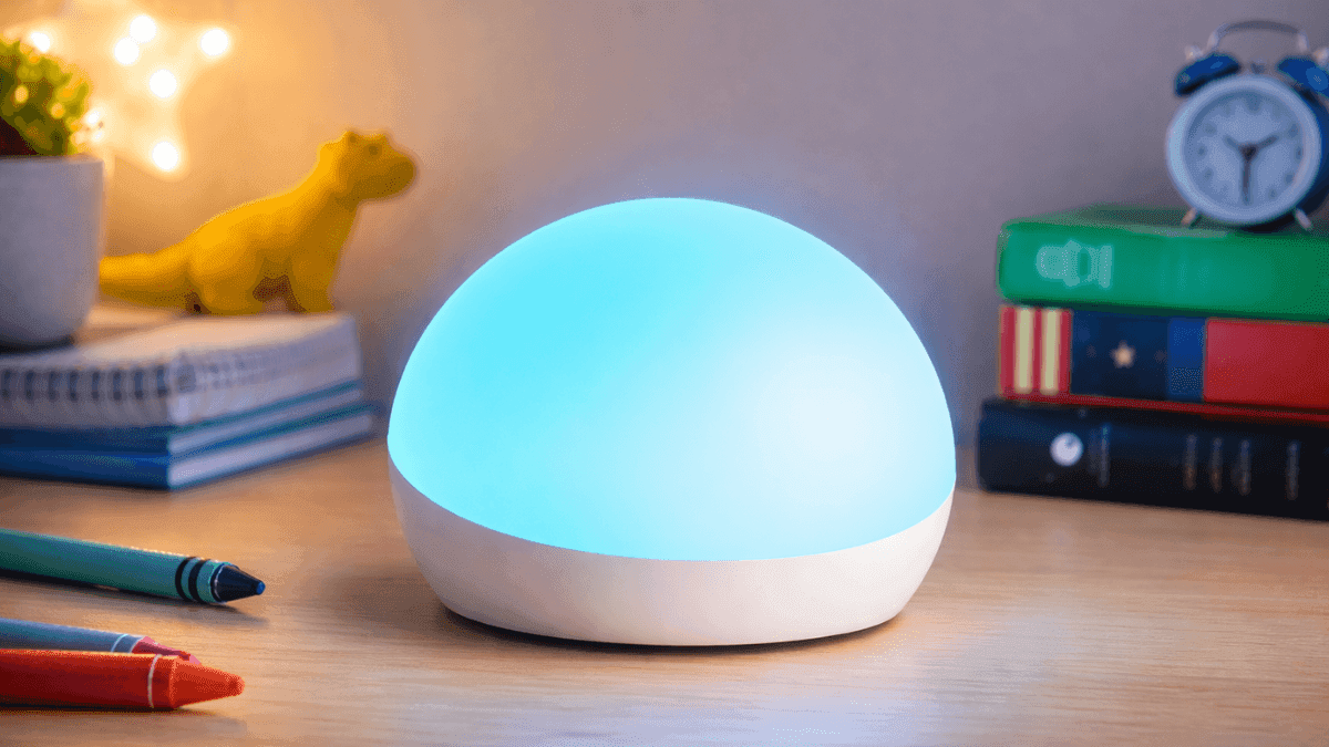 Amazon Echo Glow smart lamp glowing blue on a desk in a kids bedroom with books, crayons, and toys nearby.