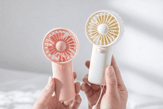 Mini portable handheld fans in pastel pink and cream colors, held in hands, compact battery-powered personal cooling devices with simple button controls and circular blade guards.