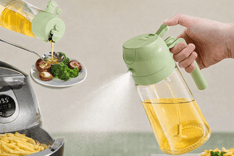 2-in-1 oil sprayer bottle for cooking with green handle, spraying fine mist of oil over food while also pouring oil onto vegetables, shown next to an air fryer and plated broccoli and mushrooms.