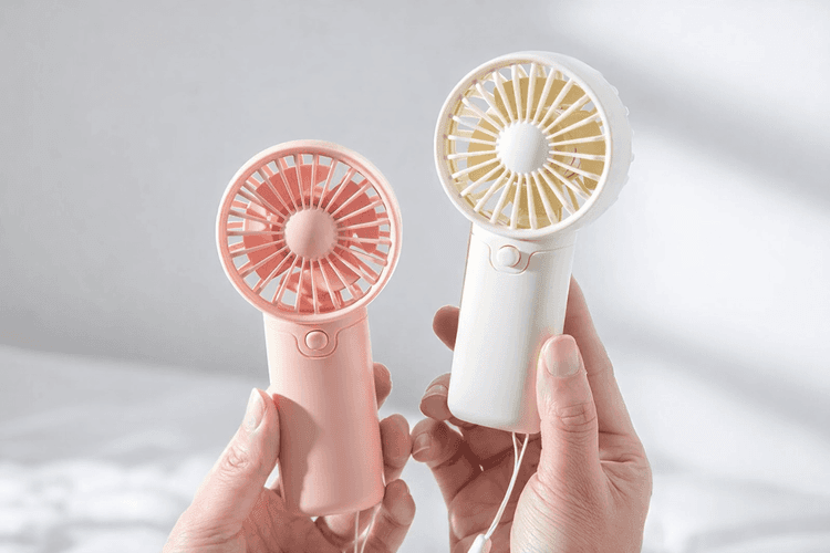 Mini portable handheld fans in pastel pink and cream colors, held in hands, compact battery-powered personal cooling devices with simple button controls and circular blade guards.